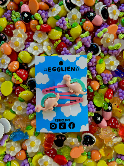 Two peach dinosaurs on bright pink hair-clips on an 'EGGLIEN' card in the center surrounded by colorful decorative items
