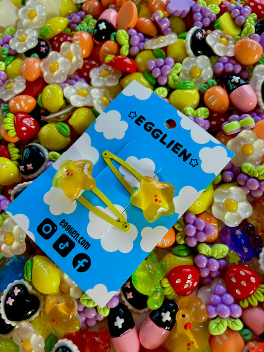 Two yellow star with happy faces hair clips on a blue card with 'EGGLIEN' branding, surrounded by colorful decorative resin pieces