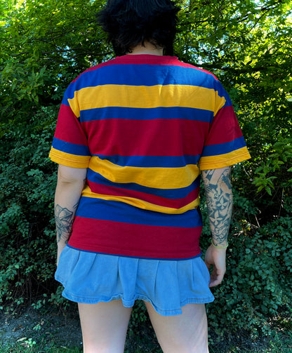 Person wearing a red, yellow, and blue striped shirt with a blue skirt outdoors with back facing the camera showing the back of the shirt