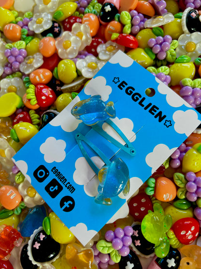 Two blue fish hair clips on a blue card with 'EGGLIEN' branding, surrounded by colorful decorative resin pieces