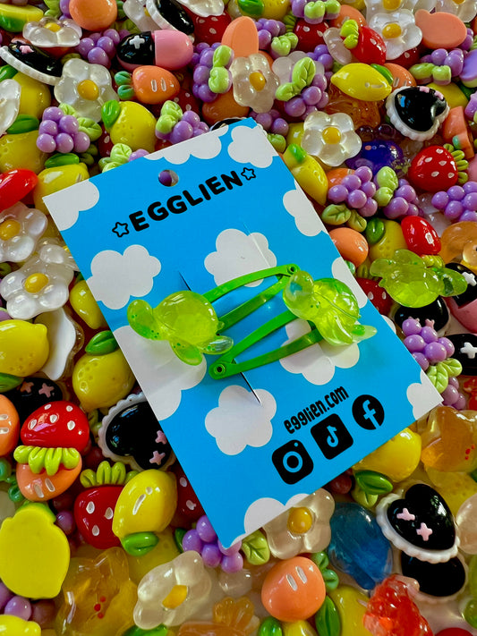 Two green turtle hair clips on a blue card with 'EGGLIEN' branding, surrounded by colorful decorative resin pieces