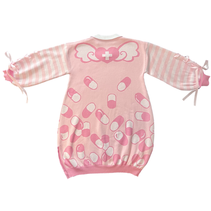 Flat lay image of the back of the pink menhera sweater dress. The sweater dress body is pastel pink with darker pink and white capsules on it. On the top part of the body there is a pink heart with a white cross that has wings coming out of the heart. The sleeves are pastel pink and white striped with pastel pink ribbons installed to form a bow on the end of the sleeves. 