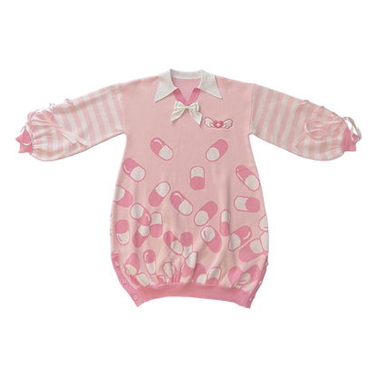 Flat lay image of the front of the pink menhera sweater dress. The sweater dress body is pastel pink with darker pink and white capsules on it. It has a white pointed collar and a removable white ribbon on the center collar. The sleeves are pastel pink and white striped with pastel pink ribbons installed to form a bow on the end of the sleeves. There is a darker pink and white embroidered heart with a cross and wings on the front left chest.