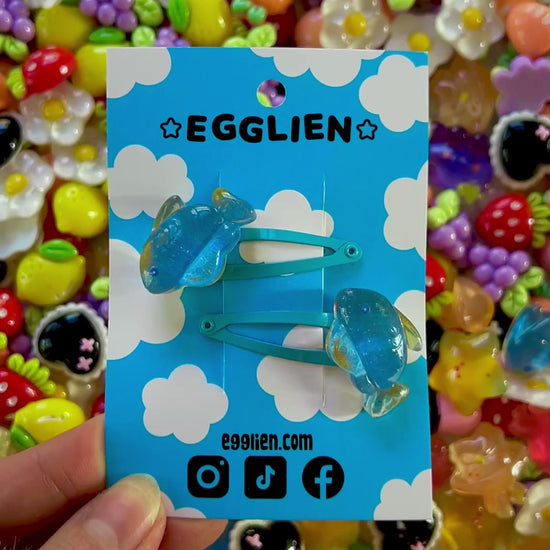 Two blue fish hair clips on a blue card with 'EGGLIEN' branding, surrounded by colorful decorative resin pieces