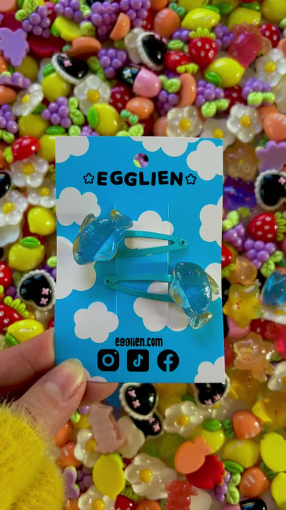 Two blue fish hair clips on a blue card with 'EGGLIEN' branding, surrounded by colorful decorative resin pieces