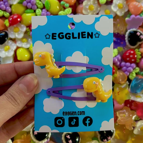 Yellow dinosaur-shaped hair clips on a blue card with 'EGGLIEN' branding, surrounded by colorful decorative resin pieces.