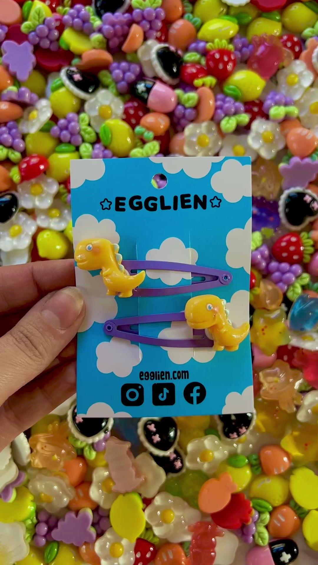 Yellow dinosaur-shaped hair clips on a blue card with 'EGGLIEN' branding, surrounded by colorful decorative resin pieces.