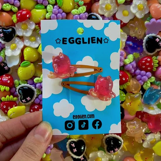 Two red/orange cuttlefish hair clips on a blue card with 'EGGLIEN' branding, surrounded by colorful decorative resin pieces