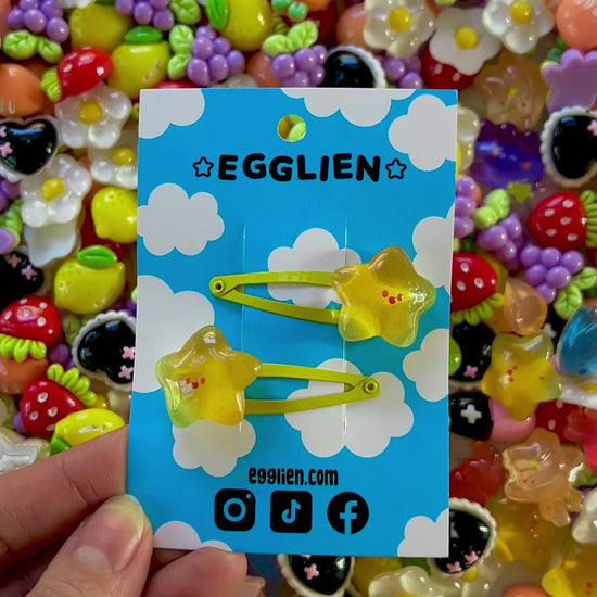 Two yellow star with happy faces hair clips on a blue card with 'EGGLIEN' branding, surrounded by colorful decorative resin pieces