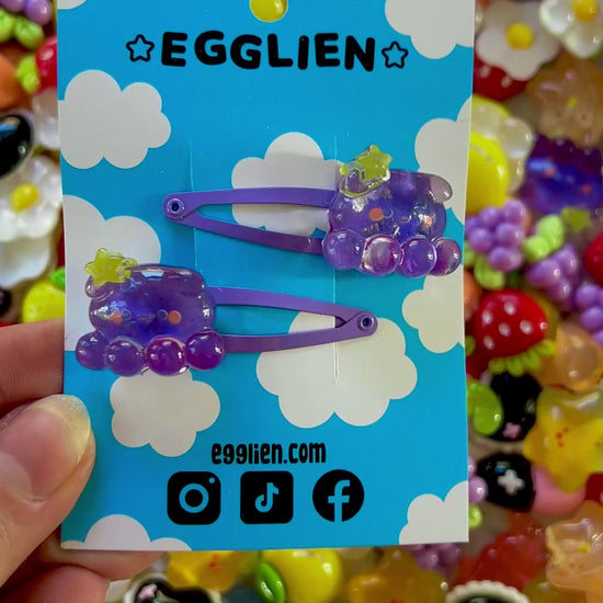 Two purple octopus  with a yellow star hair clips on a blue card with 'EGGLIEN' branding, surrounded by colorful decorative resin pieces.