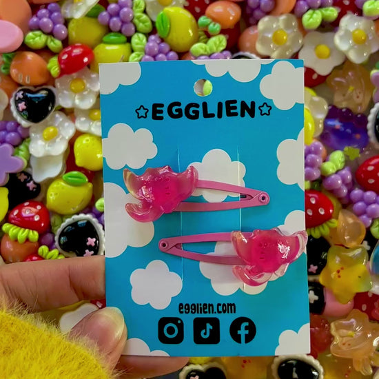 Two pink ray hair clips on a blue card with 'EGGLIEN' branding, surrounded by colorful decorative resin pieces