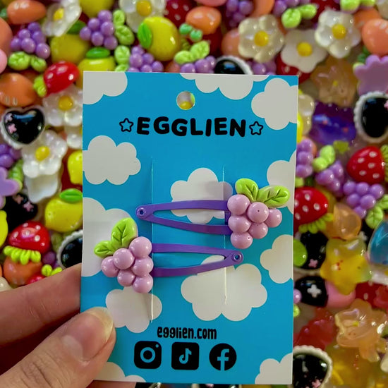 Two purple grape hair clips on a blue card with 'EGGLIEN' branding, surrounded by colorful decorative resin pieces.