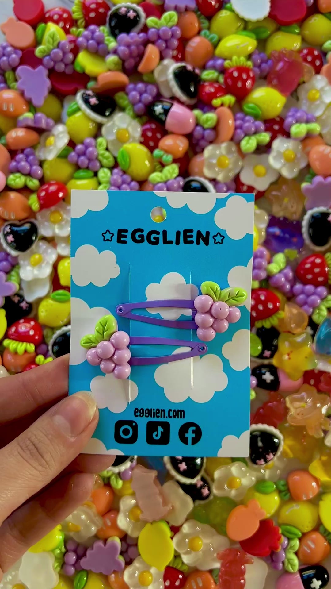 Two purple grape hair clips on a blue card with 'EGGLIEN' branding, surrounded by colorful decorative resin pieces.