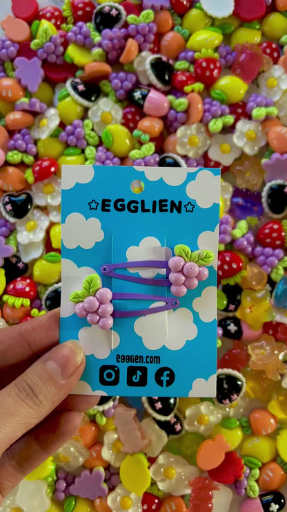 Two purple grape hair clips on a blue card with 'EGGLIEN' branding, surrounded by colorful decorative resin pieces.