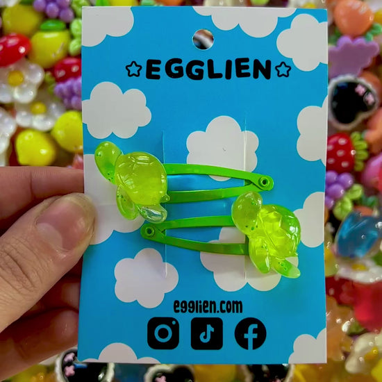 Two green turtle hair clips on a blue card with 'EGGLIEN' branding, surrounded by colorful decorative resin pieces