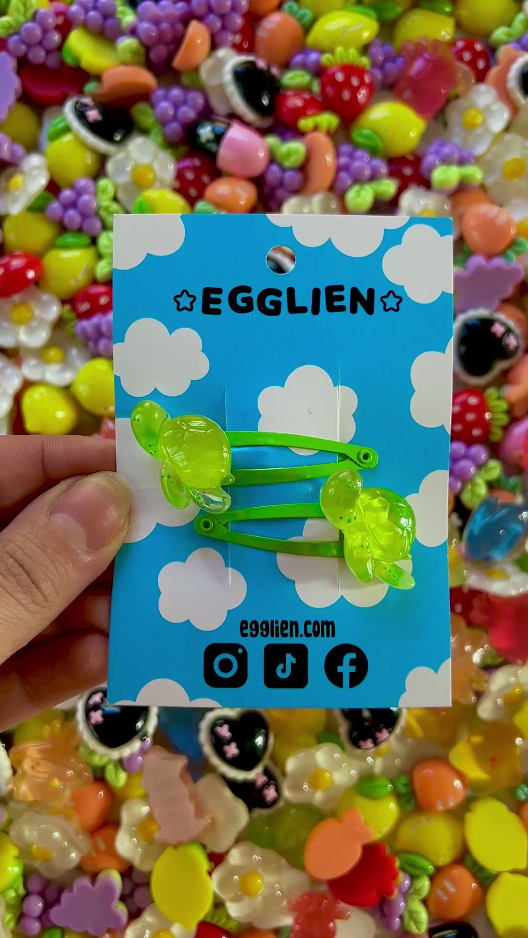 Two green turtle hair clips on a blue card with 'EGGLIEN' branding, surrounded by colorful decorative resin pieces