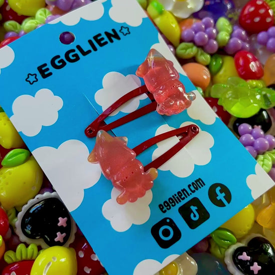 Two red squid hair clips on a blue card with 'EGGLIEN' branding, surrounded by colorful decorative resin pieces