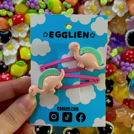 Two peach dinosaurs on bright pink hair-clips on an 'EGGLIEN' card in the center surrounded by colorful decorative items