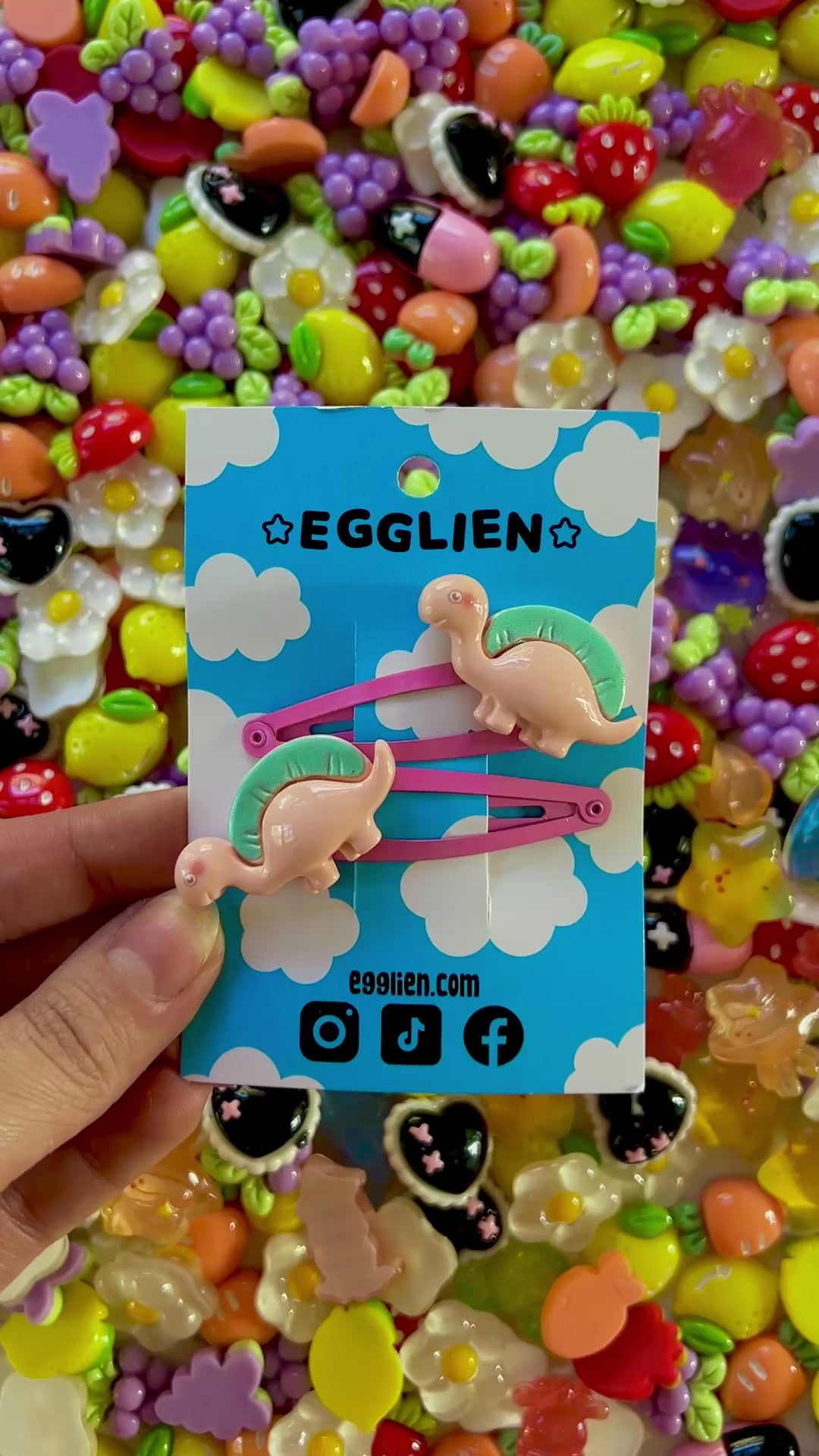 Two peach dinosaurs on bright pink hair-clips on an 'EGGLIEN' card in the center surrounded by colorful decorative items