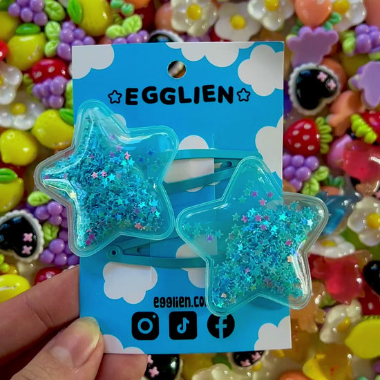 Two blue pvc stars filled with star glitter hair clips on a blue card with 'EGGLIEN' branding, surrounded by colorful decorative resin pieces