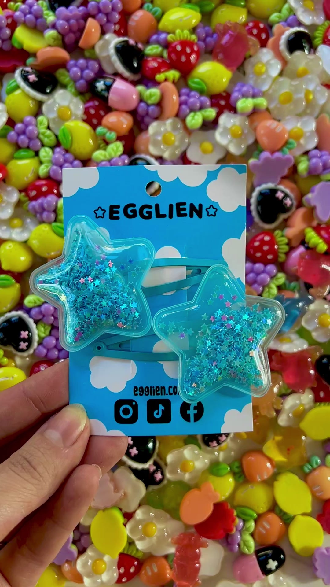 Two blue pvc stars filled with star glitter hair clips on a blue card with 'EGGLIEN' branding, surrounded by colorful decorative resin pieces