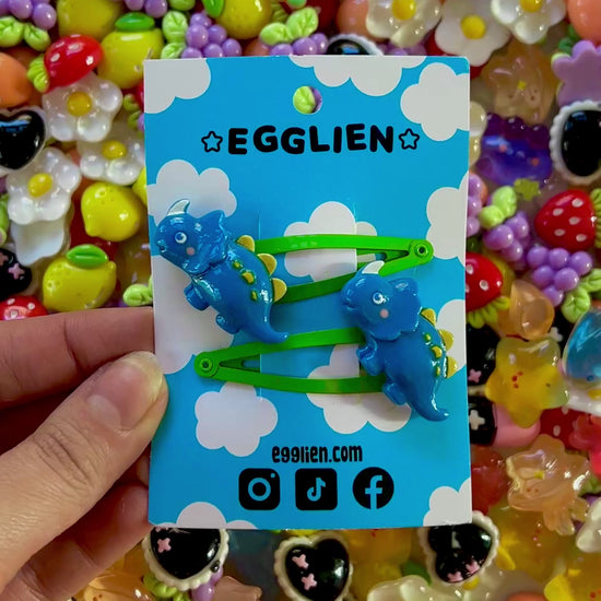 Dark Blue dinosaur-shaped hair clips on a blue card with 'EGGLIEN' branding, surrounded by colorful decorative resin pieces.
