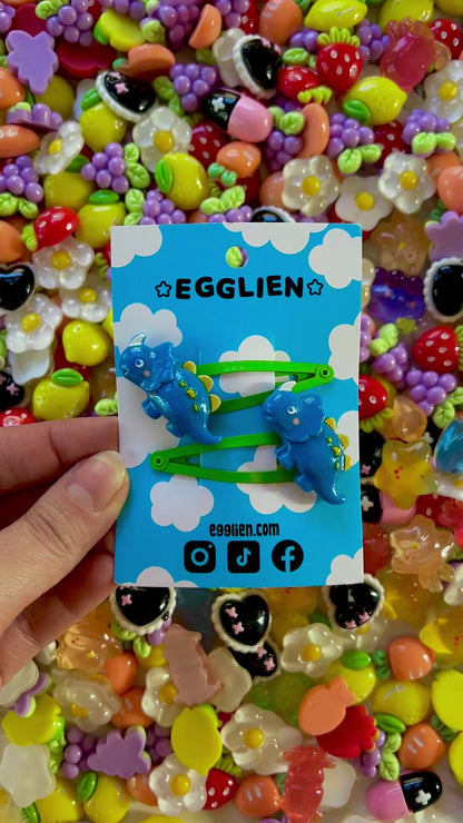 Dark Blue dinosaur-shaped hair clips on a blue card with 'EGGLIEN' branding, surrounded by colorful decorative resin pieces.