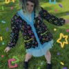A person in a green field wearing a multicolored cardigan and blue dress with colorful confetti graphics overlaid on the image. Cardigan pattern is bright yellow, pink, purple green and blue stars, circles, squares, and confetti on black background.