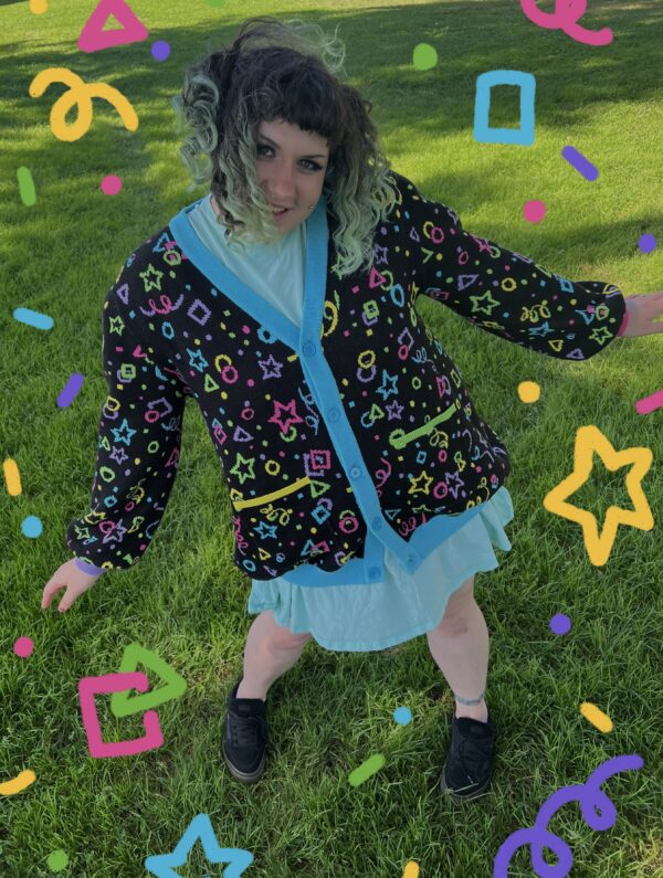 A person in a green field wearing a multicolored cardigan and blue dress with colorful confetti graphics overlaid on the image. Cardigan pattern is bright yellow, pink, purple green and blue stars, circles, squares, and confetti on black background.