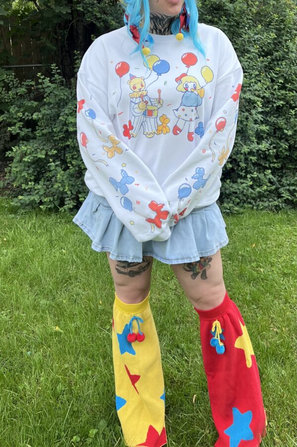 Woman standing outside wearing a white crew neck with clown themed characters, a denim skirt, and knee-high legwarmers with star patterns.