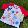 A playful red and white crop top with clown and balloon graphics and matching blue award ribbon and a sticker sheet all laying on green grass