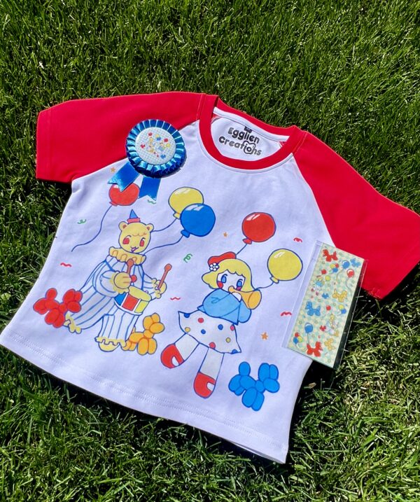 A playful red and white crop top with clown and balloon graphics and matching blue award ribbon and a sticker sheet all laying on green grass