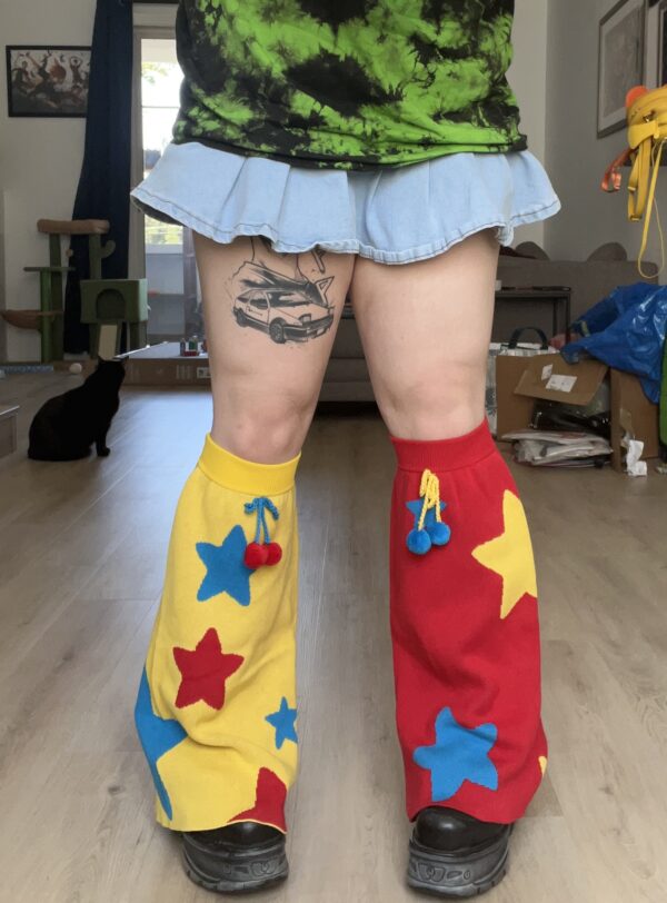 Person wearing a green and black tie dye shirt, jean skirt and colorful red and yellow legwarmers with a black cat and box in the background. Leg Warmer on the right is red with yellow and blue stars and a bow with pom poms. The leg warmer on the left is yellow with red and blue stars and a bow with pom poms.