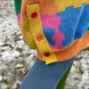 Close up image of a person wearing colorful knit design and a blue and green tights. We see the bottom orange ribbing of the dress with a yellow side cuff. The side cuff shows two red buttons and an orange button.