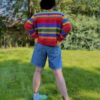 Person wearing a colorful striped sweater and denim shorts standing in a grassy area with trees.