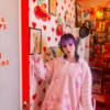 Girl is standing in room with one hand resting against the wall. She is wearing a pastel pink sweater dress with darker pink and white capsules on it with a pastel pink and white striped sleeves. Pastel pink ribbons are peaking out from the sleeves. In the background is room with cherry shelves, red heart wallpaper and pictures of women on the wall.