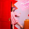 Girl is playfully opening a red door with three quarters of her body peaking out. She is wearing a pastel pink sweater dress with darker pink and white capsules on it with a pastel pink and white striped sleeves. In the background is picture of a closed eye showing eyelashes and an LED sign that reads "Sorry Daddy.. I'm In.." half of the sign is cut off.