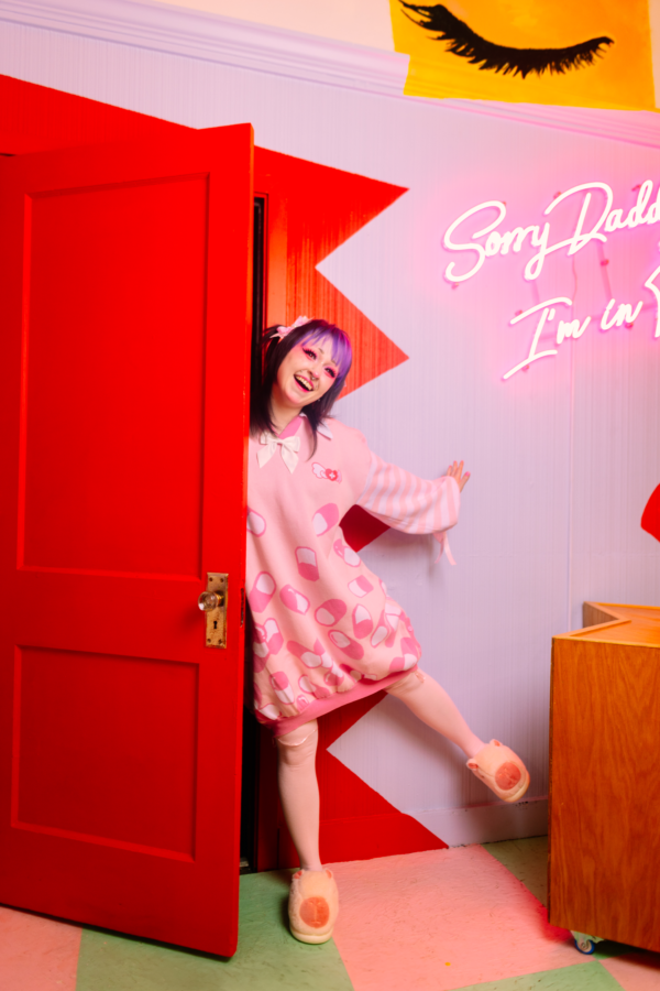 Girl is playfully opening a red door with three quarters of her body peaking out. She is wearing a pastel pink sweater dress with darker pink and white capsules on it with a pastel pink and white striped sleeves. In the background is picture of a closed eye showing eyelashes and an LED sign that reads "Sorry Daddy.. I'm In.." half of the sign is cut off.