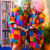 Two girls are standing in the center. One girl is wispering to the other while she reads. Both are wearing colorful sweater dresses that have the design of puzzle play mats on them. Behind them is a bookshelf filled with books, a plant, and a large sculpture of a woman.