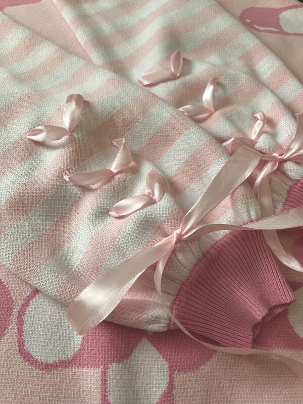 The pastel pink and white sleeves of a knit cardigan are shown close up. Showcasing the darker pink cuffs along with the pastel pink ribbons that are installed in each of the sleeves. The ribbon is shaped in three hearts that get closer to the cuffs where they combine to create a big pastel pink bow.