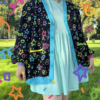 A girl in a green field wearing a multicolored cardigan and blue dress with colorful confetti graphics overlaid on the image. Cardigan pattern is bright yellow, pink, purple green and blue stars, circles, squares, and confetti on black background.