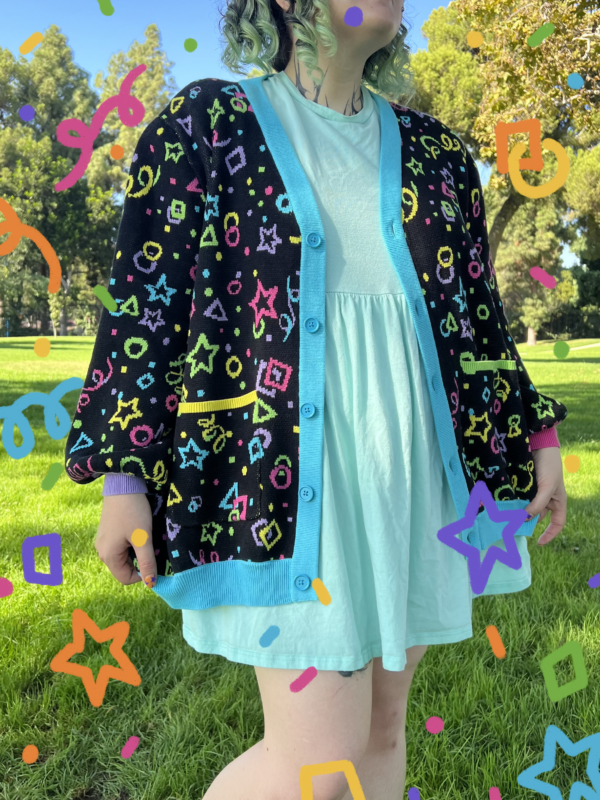 A girl in a green field wearing a multicolored cardigan and blue dress with colorful confetti graphics overlaid on the image. Cardigan pattern is bright yellow, pink, purple green and blue stars, circles, squares, and confetti on black background.