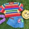 Rainbow striped sweater, purple slip on shoes, happy face backpack, red sunglasses, jean shorts with two plush keychains attached all laying flat on green grass