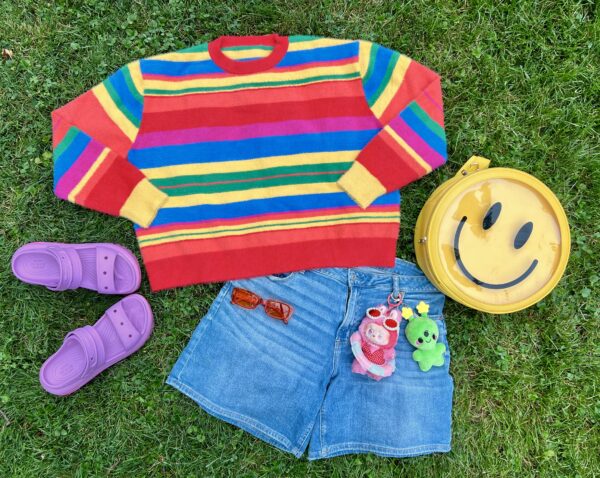 Rainbow striped sweater, purple slip on shoes, happy face backpack, red sunglasses, jean shorts with two plush keychains attached all laying flat on green grass