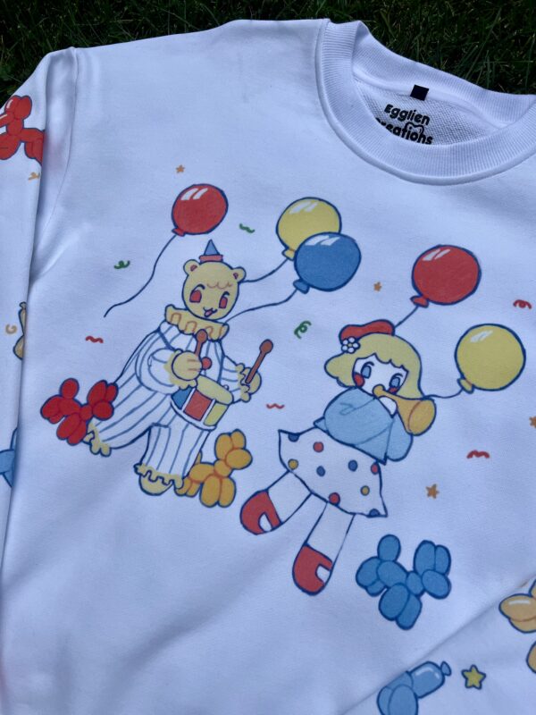 Close up photo of a white crew neck with clown and balloon graphics laying on grass