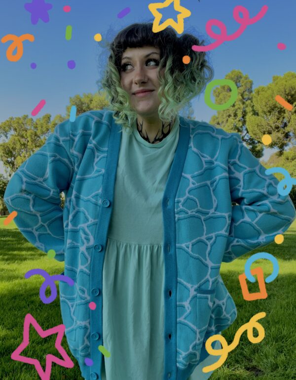 A person standing on grass wearing a blue cardigan with a geometric water pattern, paired with a lighter blue dress. There are colorful confetti-like shapes overlaid on the image.