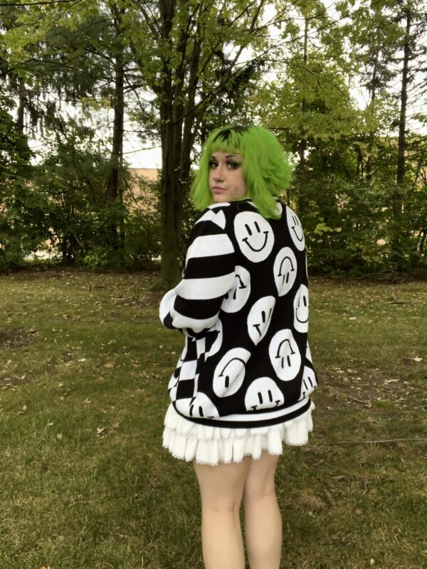 Girl standing in a green field with back facing the camera wearing a black and white happy face cardigan and a white skirt.