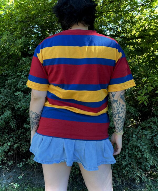Person wearing a red, yellow, and blue striped shirt with a blue skirt outdoors with back facing the camera showing the back of the shirt