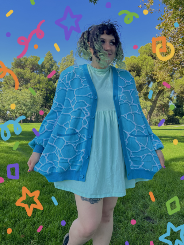 A person standing on grass wearing a blue cardigan with a geometric water pattern, paired with a lighter blue dress. There are colorful confetti-like shapes overlaid on the image.