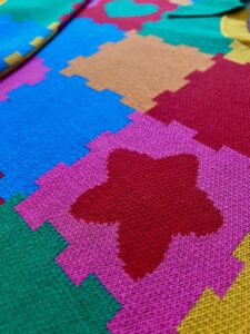 Colorful knit sweater dress is laying on the ground.The dress is bright blue, green, orange, pink, yellow and red with the design of puzzle playmats. Close up we see the texture of the knitted fibers.